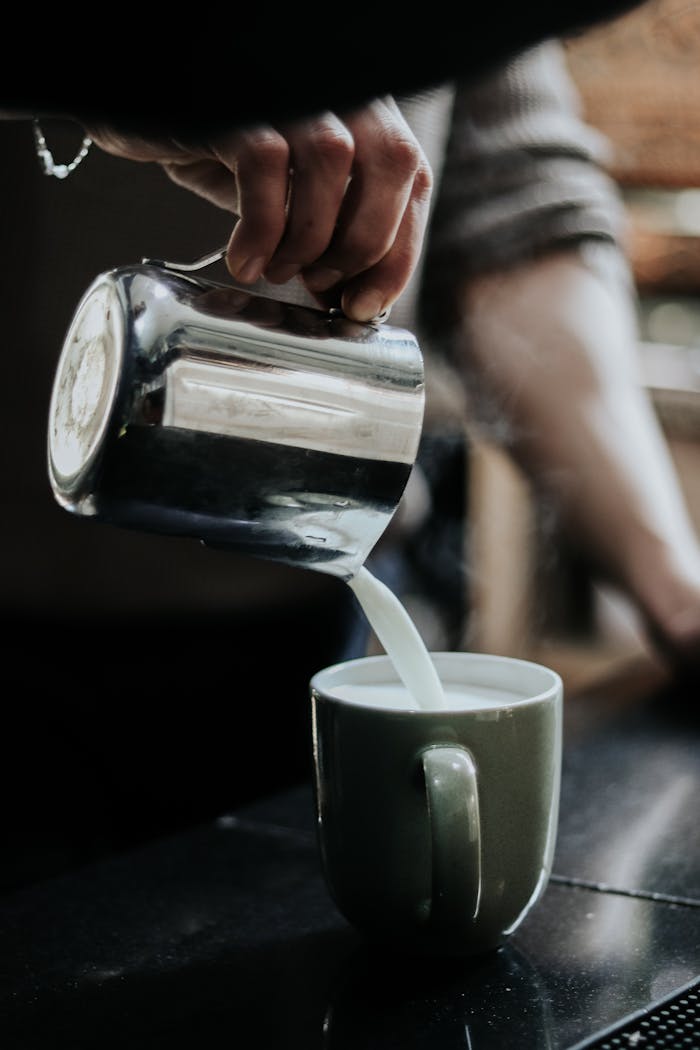 A person pours steaming milk into a cappuccino mug, creating a warm beverage.